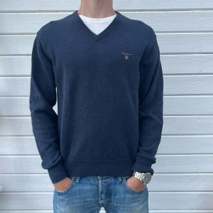 Blå v-ringad tröja från GANT - Mörkblå v-ringad tröja från GANT med broderad logga på bröstet. Tröjan är i mjuk bomull, har ribbade muddar och lång ärm. Perfekt för dig som gillar en clean och klassisk stil.