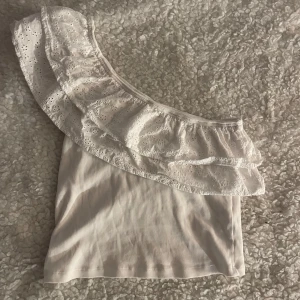 Vit oneshoulder topp med volang Gina Tricot - Supersöt vit oneshoulder topp från Gina Tricot med bred volangkant i broderad spets. Ribbad struktur i tyget och ärmlös design. Perfekt för dig som vill sticka ut med en feminin och trendig look.