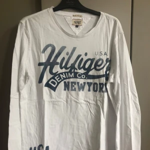 Vit långärmad Hilfiger Denim tröja - Vit långärmad tröja från Hilfiger Denim med stort blått tryck på bröstet där det står 'Hilfiger Denim Co. New York'. Tröjan har rund halsringning och är gjord i mjuk bomull. Perfekt för en chill och stilren look.