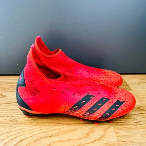 Röda Adidas Predator fotbollsskor - Säljer ett par HELT NYA röda Adidas Predator fotbollsskor med svarta detaljer och ikoniska tre ränder på sidan. Skorna har en stickad ovandel och gummitaggar för extra grepp på bollen. Sulan är designad för bra fäste på både gräs och konstgräs. Perfekt för dig som vill sticka ut på planen.
