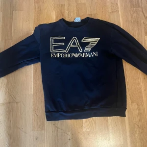 Svart sweatshirt EA7 Emporio Armani - Svart sweatshirt från EA7 Emporio Armani med stort guldigt logotryck på bröstet. Klassisk rund halsringning och långa ärmar. Mjuk och skön bomullsblandning, perfekt för en stilren och sportig look.