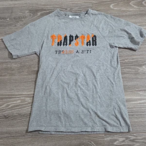 Grå Trapstar t-shirt - Snygg grå t-shirt från Trapstar med fet logga i svart och orange på bröstet. Klassisk passform och korta ärmar, perfekt till jeans eller shorts. Mjuk bomullskänsla och street vibe.