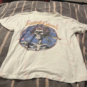 Grateful Dead t-shirt med tryck - Vit t-shirt med färgstarkt Grateful Dead-tryck framtill. Motivet har en dödskalle med rosor och blå bakgrund. Klassisk passform och korta ärmar, tillverkad i mjuk bomull. Perfekt för dig som gillar vintageband och coola prints.