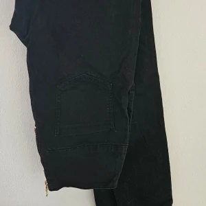 Svarta cargopants från Esmara - Svarta cargopants från Esmara med flera fickor och snygga guldfärgade dragkedjor vid benen. Byxorna har en loose passform och är tillverkade i mjukt bomullstyg. Perfekta för en avslappnad och cool streetstil.