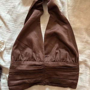 Brun halterneck topp från Bik Bok - Snygg brun halterneck topp från Bik Bok i glansigt material. Toppen har djup v-ringning, breda axelband och veckade detaljer i midjan. Stängs med små hyskor i ryggen. Perfekt för dig som vill sticka ut med en trendig look.
