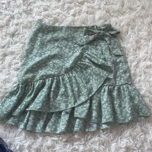 Grön blommig omlottkjol med volang - Supersöt ljusgrön kortkjol med vitt blommönster och volangkant. Kjolen har omlottmodell med knytband i midjan som ger en extra gullig touch. Perfekt för vår och sommar när du vill ha en fräsch och feminin look.