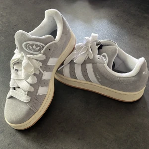 Grå Adidas Campus sneakers i mocka - Snygga grå Adidas Campus sneakers i mjuk mocka med klassiska vita ränder och vit sula. Skorna har rund tå, platt sula och breda vita skosnören. Perfekta för en clean och stilren streetlook.