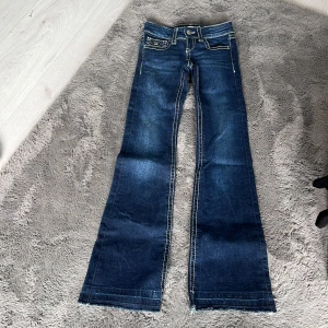 Blå bootcut jeans Gina Tricot 134 - Snygga blå jeans från Gina Tricot i storlek 134. Modellen har bootcut-ben, markerade kontrastsömmar och klassiska fickor med lock och knapp bak. Jeansen är i stretchigt denim och har låg midja. Perfekta för dig som gillar retrovibbar och cool stil.