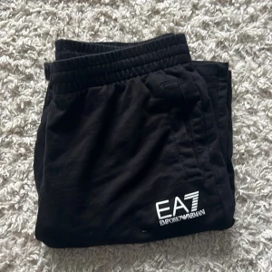 Svarta EA7 Emporio Armani mjukisbyxor - Svarta mjukisbyxor från EA7 Emporio Armani med vit logga på benet. Byxorna har resår i midjan och är tillverkade i mjukt material som passar perfekt för chill eller träning. Snygg och enkel design med fickor på sidorna.