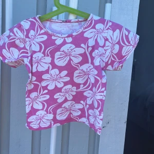 Rosa blommig t-shirt med kort ärm - Snygg rosa t-shirt med stora vita blommor över hela tyget. T-shirten har rund halsringning och korta ärmar, perfekt för varma dagar. Skön och mjuk känsla, passar dig som gillar färg och mönster.