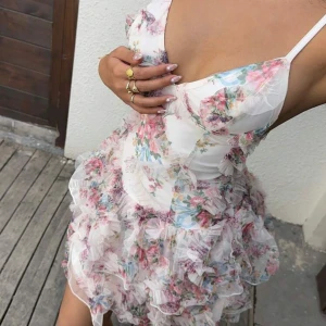 Blommig volangklänning med v-ringning - Supersöt kort klänning med blommigt mönster i rosa, blått och grönt på vit botten. Djup v-ringning, smala axelband och flera lager volanger i tyll för en fluffig look. Tryck på KÖP NU så postar jag så snabbt som möjligt!! 🩷🩷