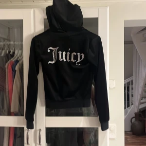 Svart Juicy Couture velourjacka XXS - Svart velourjacka från Juicy Couture med huva, dragkedja och fickor fram. Stora glittriga 'Juicy'-bokstäver på ryggen och JC-logga på bröstet. Snygg croppad passform och klassiska detaljer som snörning i huvan och ribbade muddar.
