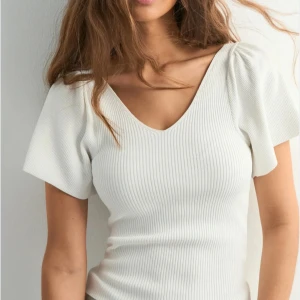 Vit ribbad topp med knyt i ryggen - Trendig Supersöt vit ribbad topp med v-ringning både fram och bak. Korta, lite puffiga ärmar och en fin knyt-detalj i ryggen. Sitter snyggt och har en feminin siluett. Perfekt för dig som gillar stilrena och trendiga plagg med det lilla extra. Finns minimalt med täcken att den har används. Ny pris 350 mitt pris 180 kr