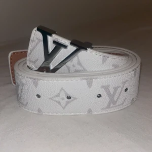 Vit Louis Vuitton monogram skärp - Säljer ett vitt Louis Vuitton skärp i skinn med klassiskt monogrammönster i ljusgrått och stor LV-logga i silvermetall som spänne. Bältet har en stilren och lyxig vibe, perfekt för att lyfta vilken outfit som helst.