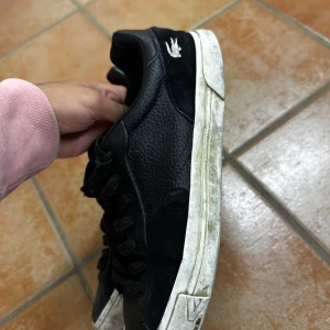 Svarta sneakers från Lacoste - Svarta Lacoste sneakers med klassisk krokodillogga på hälen. Skorna har en mix av slätt och texturerat skinn samt detaljer i mocka. De har svarta snören och vit platt sula för en clean look. Perfekta för dig som gillar stilrena och sportiga vibes.