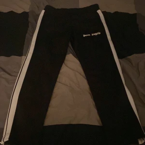 Palm Angels svarta mjukisbyxor storlek L - Svarta mjukisbyxor från Palm Angels med vita sidoränder och logga på låret. Byxorna har en sportig vibe och är perfekta för en chill och trendig look. Elastisk midja och raka ben ger en skön passform.