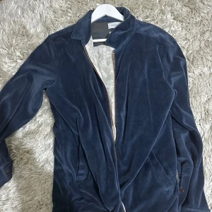 Blå sammet bomberjacka Limitato - Snygg blå bomberjacka i mjuk sammet från Limitato. Jackan har dragkedja framtill, klassisk krage och ett unikt tryck på ryggen med texten 'What if this is all real?'. Perfekt för dig som vill sticka ut med stilrena detaljer och coolt motiv.