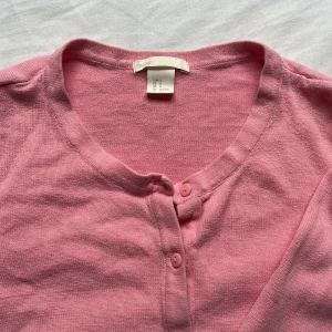 Rosa kofta från H&M Basic, S - Söt rosa kofta från H&M Basic i storlek S. Koftan har rund halsringning, långa ärmar och knäppning framtill med matchande rosa knappar. Perfekt för lager-på-lager och enkel att styla till olika outfits.