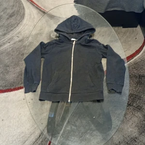 Svart hoodie med dragkedja från 157 - En svart hoodie från 157 i storlek 140. Tröjan har huva, långa ärmar och hel dragkedja framtill. Två fickor på magen och mjukt material som känns skönt mot huden. Perfekt för en chill och avslappnad stil.