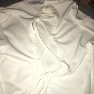 Vit ribbad polotröja med snörning - Säljer en vit ribbad polotröja med lång ärm och snygg snörningsdetalj i ryggen. Tröjan har en hög krage och är i ett stretchigt material som sitter snyggt på kroppen. Perfekt för dig som gillar trendiga detaljer och vill sticka ut lite extra.