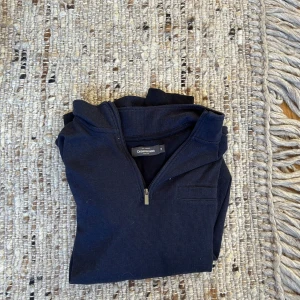 Mörkblå half zip tröja Dressmann - Mörkblå långärmad tröja från Dressmann med half zip-dragkedja och liten bröstficka. Tröjan har en klassisk krage och är tillverkad i ett mjukt bomullsmaterial. Perfekt för lager-på-lager och enkel att matcha med jeans eller chinos.