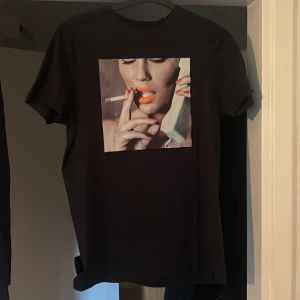 Svart Limitato t-shirt med tryck - Svart t-shirt från Limitato i 100% merceriserad bomull. Framsidan har ett stort färgstarkt tryck av en kvinna med cigarett och telefon. Klassisk passform och korta ärmar, perfekt för dig som vill sticka ut med en unik look.