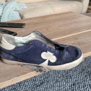 Blå mockasneakers med vit detalj - Snygga sneakers i blå mocka med vit sula och en stor vit klöverdetalj på sidan. Skon har rund tå och är utan snörning. Insidan är ljus och hälen har en vit panel. Perfekt för dig som gillar unika detaljer och streetstyle.