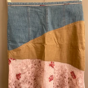 Patchwork kjol i jeans, manchester och blommigt - Unik långkjol med cool patchwork-design! Överdel i blått jeans, mittenparti i beige manchester och nederdel i ljusrosa tyg med blommigt mönster i rött och brunt. Kjolen har dragkedja bak och är tillverkad i bomull och polyester. Perfekt för dig som gillar retro och mixade material. Midjemått 76 cm , längd 50 cm