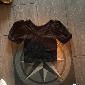 Svart croppad topp med puffärm - Svart croppad topp med korta puffärmar och rund halsringning. Tyget är strukturerat och ger en cool känsla. Perfekt för dig som gillar trendiga detaljer och vill sticka ut lite extra.