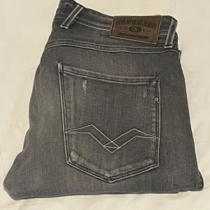Replay Anbass grå jeans med slitningar - Snygga Replay Anbass jeans i grå tvätt med slitna detaljer och coola sömmar på bakfickorna. Jeansen har en slim passform och klassisk femficksdesign. Perfekta för dig som gillar en avslappnad men trendig look.