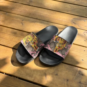 Gucci sandaler med tiger och blommor - Svarta sandaler från Gucci med bred rem i konstmaterial, dekorerad med färgglada tigrar och rosa blommor på brun bakgrund. Snygg och unik design som sticker ut, perfekt för sommaren. Sula i gummi med präglad Gucci-logga undertill.