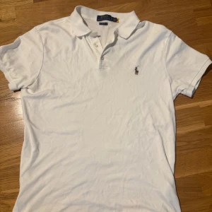 Vit pikétröja från Polo Ralph Lauren - Klassisk vit pikétröja från Polo Ralph Lauren i slim fit-modell. Tröjan har korta ärmar, krage med knappar och den ikoniska broderade loggan på bröstet. Tillverkad i mjuk bomull som känns skön mot huden. Perfekt för en clean och stilren look.