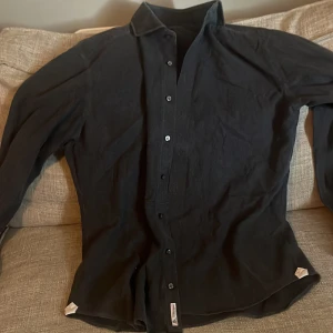 Svart skjorta från La Chemise - Stilren svart skjorta från La Chemise med slim fit-passform. Skjortan har klassisk krage, långa ärmar och knäppning framtill. Perfekt för dig som gillar en enkel och snygg look. Materialet känns lätt och passar året runt.