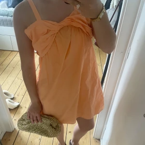 Persikofärgad kort klänning med rosett - Supersöt persikofärgad kort klänning med breda axelband och stor rosettdetalj framtill. Luftig modell med lätt A-linjeform, perfekt för varma dagar. Klänningen har en fräsch och somrig vibe och är tillverkad i ett tunt, mjukt tyg. Storlek 13-14 år men passar xs