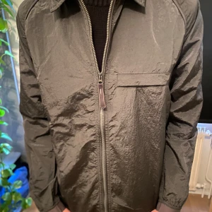 Grå vindjacka från Stone Island - Säljer en grå vindjacka från Stone Island med klassisk patch på ärmen. Jackan har dragkedja framtill, krage och en bröstficka. Perfekt för sommaren då materialet är tunt.