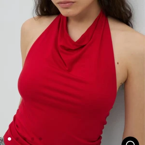 Röd halterneck topp med drapering - Snygg röd halterneck topp med draperad urringning och öppen rygg. Toppen är ärmlös och har en tight passform som framhäver figuren. Perfekt att matcha med jeans eller kjol för en cool look. Tillverkad i mjukt och stretchigt material.
