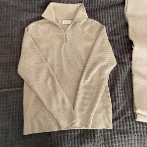 Beige ribbad half zip-tröja East West - Säljer en beige ribbstickad tröja från East West i 100% bomull. Tröjan har hög krage, half zip-dragkedja och långa ärmar. Perfekt för lager-på-lager och chill stil. Passformen är normal och materialet känns mjukt mot huden.