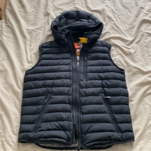 Parajumper dunväst med huva - Säljer en svart dunväst från Parajumpers i storlek S. Västen är quiltad, har huva och dragkedja framtill samt två snedställda fickor med dragkedja. Insidan är ljusgrå och västen är superlätt, perfekt för lager-på-lager. Klassisk Parajumpers-detalj vid halsen.