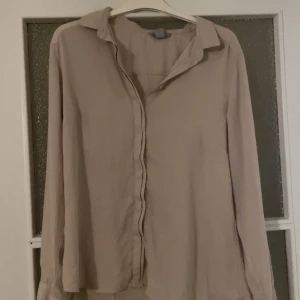 Beige långärmad skjorta - Stilren beige skjorta med klassisk krage och långa ärmar. Skjortan har en avslappnad passform och knäppning framtill. Perfekt för dig som gillar en enkel och clean look.
