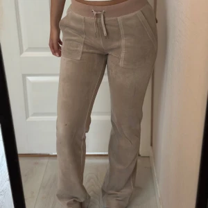 Juicy Couture mjukisbyxor - Snygga beige Juicy Couture mjukisbyxor i mjukt material med bred resår och snörning i midjan. Byxorna har raka ben, stora fickor fram och bakfickor med knappdetalj. Perfekta för en chill och avslappnad stil. 🩷