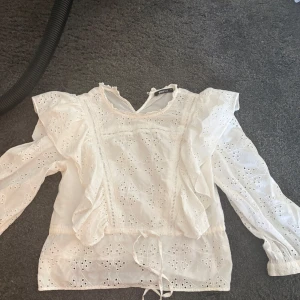 Vit broderad blus från Gina Tricot - Superfin vit blus med broderade detaljer och volanger på axlarna. Långärmad modell med knyt i midjan och vackert hålmönster över hela toppen. Perfekt för dig som gillar romantisk och luftig stil.