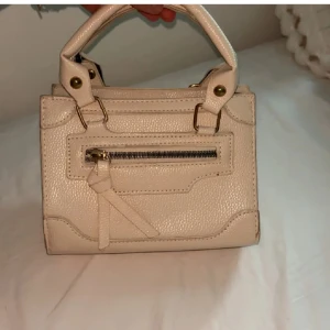 Beige handväska med dragkedja - Säljer en stilren beige handväska med guldfärgade detaljer och praktiskt handtag. Väskan har en liten ficka med dragkedja framtill och är tillverkad i ett syntetmaterial med lätt struktur. Använd ett fåtal gånger och ett band tillkommer 😊
