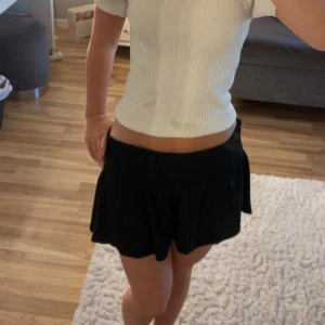 Svart kortkjol från Gina Tricot - Supersöt svart kortkjol från Gina Tricot med smockad midja och lätt volangig nederkant. Kjolen är enkel och stilren, perfekt till crop tops eller hoodies. Materialet är mjukt och stretchigt för en bekväm passform.