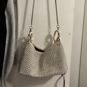 Beige axelväska med silverpaljetter - Snygg beige axelväska med massor av glittrande silverpaljetter och en cool kedjerem i metall. Väskan har en mjuk form och detaljer i ljus plast. Perfekt för att lyfta vilken outfit som helst.