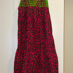 Färgstark långklänning med mönster - Säljer en midi-klänning med grön smockad överdel och färgstark rosa kjol med unikt svart och gult mönster. Klänningen har ärmlös design och luftig passform, perfekt för dig som gillar att sticka ut med coola färger och mönster.