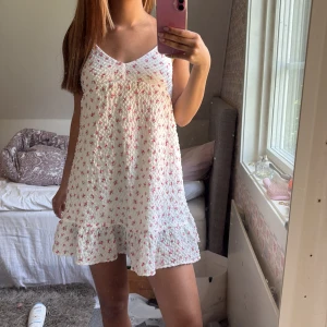 Vit blommig sommarklänning Gina Tricot - Supersöt vit kortklänning från Gina Tricot med små rosa blommor och volang nertill. Klänningen är ärmlös med smala axelband och har en lätt v-ringning. Perfekt för varma dagar och har en luftig, flowy passform.