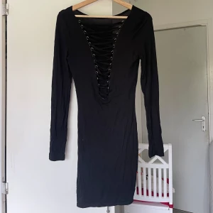 Svart kort klänning med korsettdetalj - Svart, tight klänning med lång ärm och djup v-ringning baktill med korsett snörningsdetalj. Klänningen är kort och stretchigt och har en stilren, enkel design, Man kan ha korsetten framtill eller baktill för en trendig look