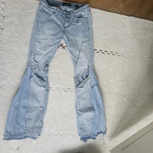 Ljusblå bootcut jeans Lucky Brand - Ljusblå jeans från Lucky Brand med bootcut passform och slitna detaljer. Jeansen har lagade partier med synliga sömmar och råa, fransiga benslut. Klassisk femficksmodell i mjukt denimtyg, perfekt för en avslappnad och edgy stil.