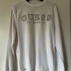 Vit långärmad tröja från Guess - Vit långärmad tröja från Guess med stor svart logga och texten 'Los Angeles' framtill. Klassisk rund halsringning och mjukt material som känns skönt mot huden. Perfekt för dig som gillar stilrena och enkla plagg med tydlig branding.