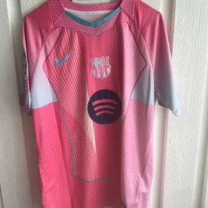 FC Barcelona Lamine Yamal rosa tröja - Snygg FC Barcelona fotbollströja i rosa och ljusblått med Lamine Yamal och nummer 10 på ryggen. Tröjan har korta ärmar, Nike-logga, klubbmärke och Spotify som sponsor framtill. Tillverkad i lätt och ventilerande material, perfekt för match eller träning.